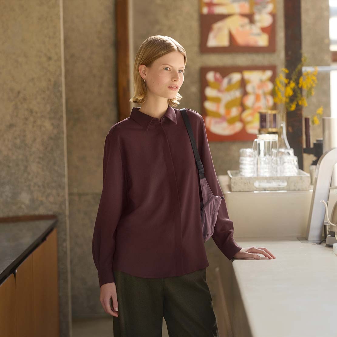 model wearing wine coloured blouse with a muted purple shoulder bag while leaning on a counter with glasses and a water jug