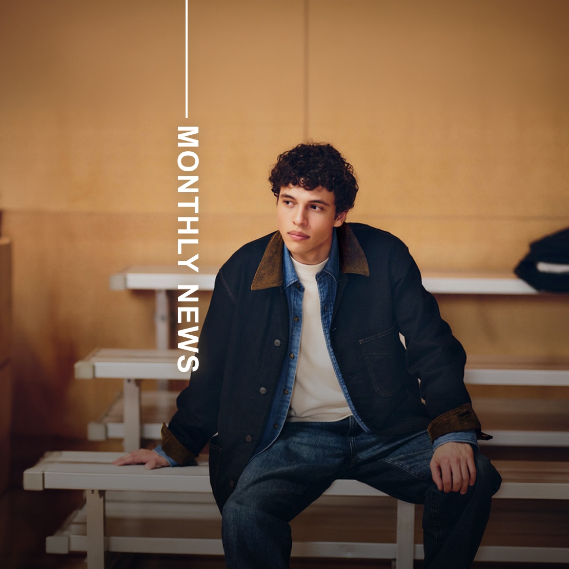 model wearing denim coverall over a mid-wash blue denim shirt and white T-shirt with mid-wash denim jeans while sat on a bench in a sports hall