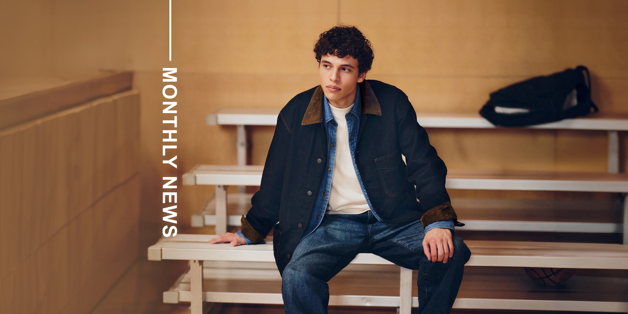 model wearing denim coverall over a denim shirt and white T-shirt with mid-wash denim jeans while sitting in a sports hall next to the words monthly news