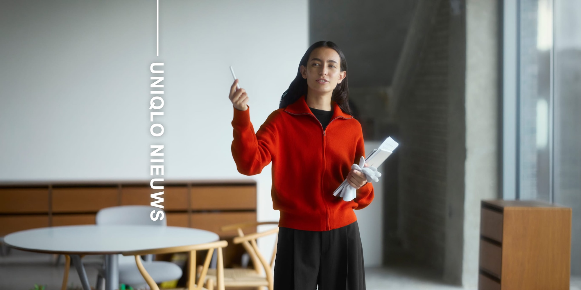 Model draagt ​​een rood vest met rits over een zwarte top met een donkerbruine broek en geeft een presentatie naast de woorden 'maandelijks nieuws'.