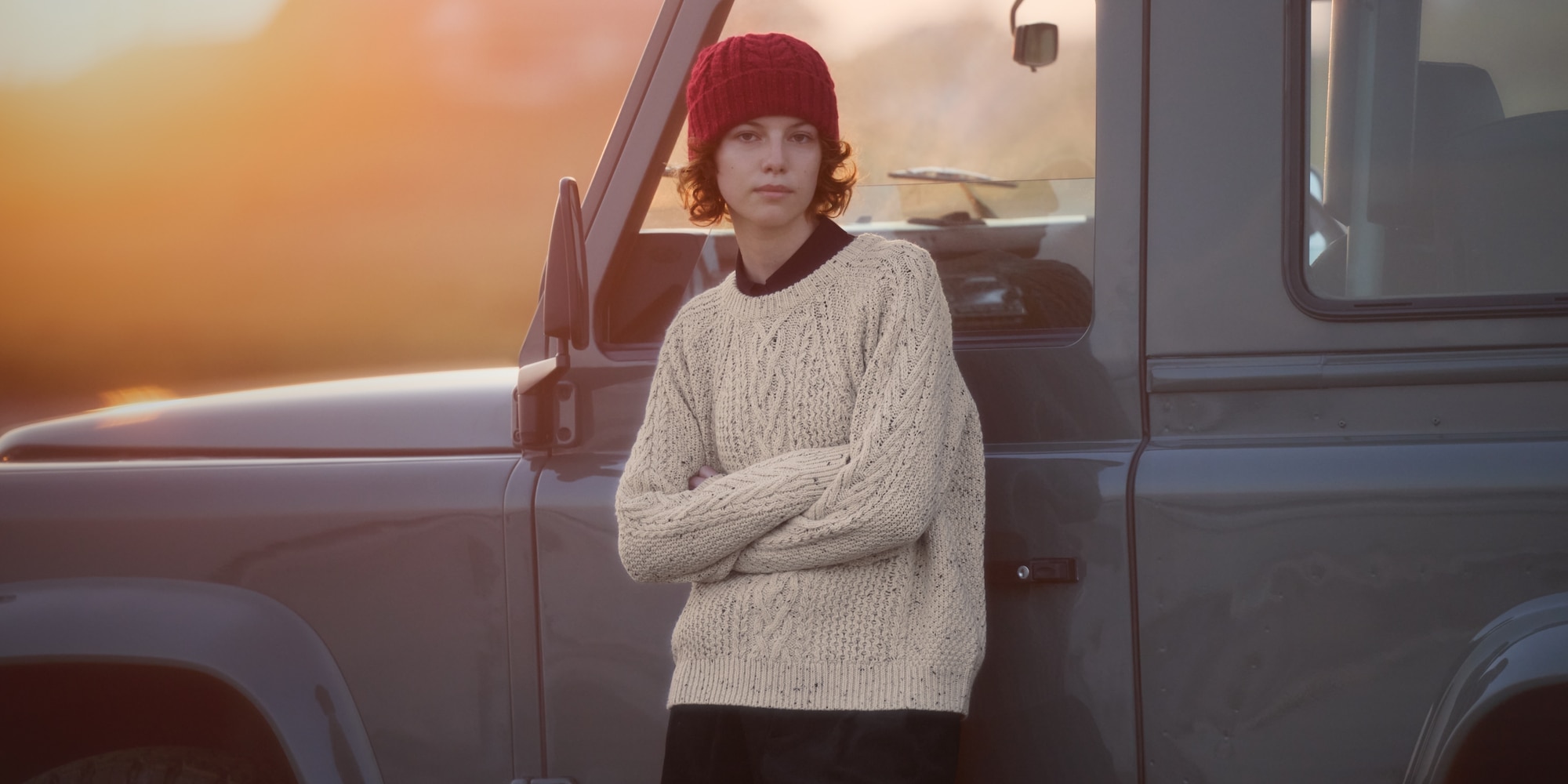 model wearing a cream cable knit jumper with a red cable knit beanie while leaning against the side of a truck with a sunset in the background