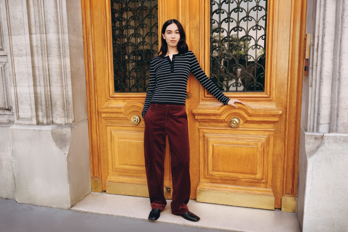model wearing navy and white striped henley with dark red trousers and black loafers while leaning against a large wooden door
