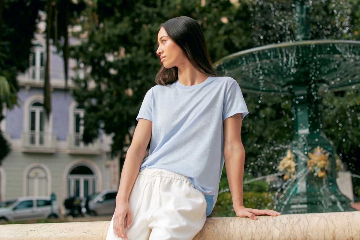model wearing light blue T-Shirt and off white trousers leaning against the side of a fountain