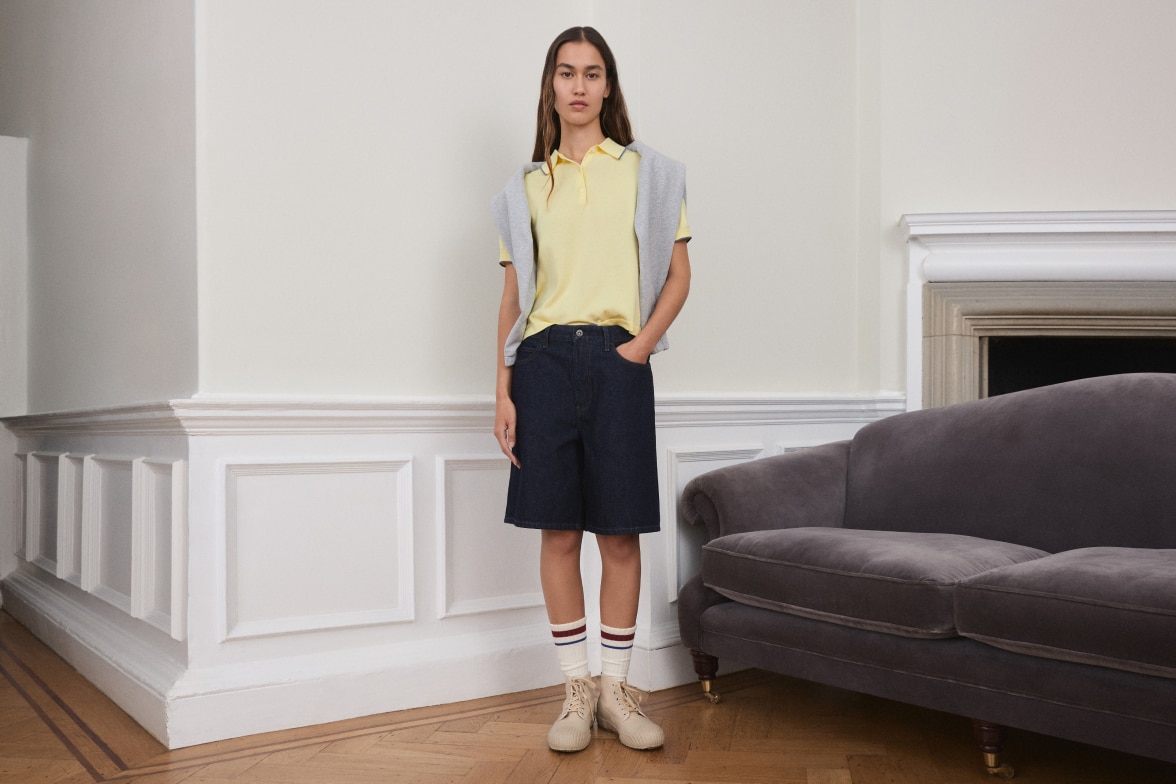model wearing denim shorts with a yellow polo shirt and grey sweatshirt draped over the shoulders
