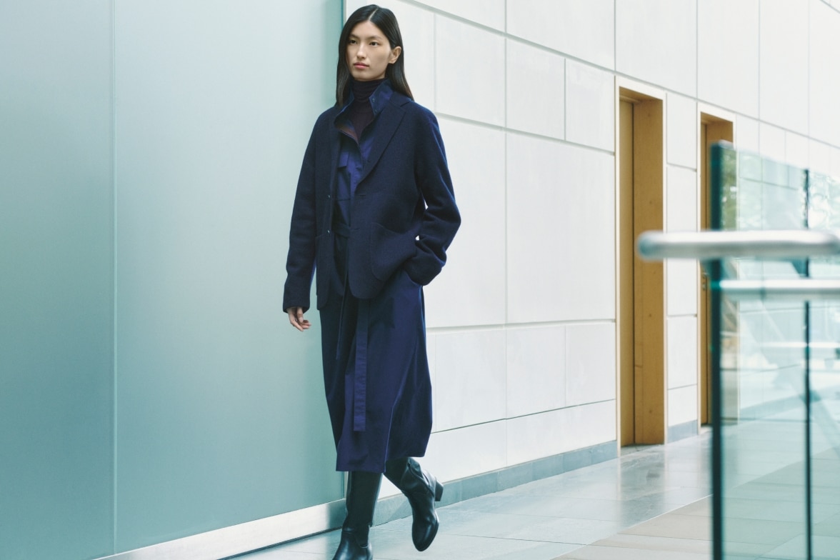 model wearing dark blue dress under a blazer with knee high boots while walking down a corridor