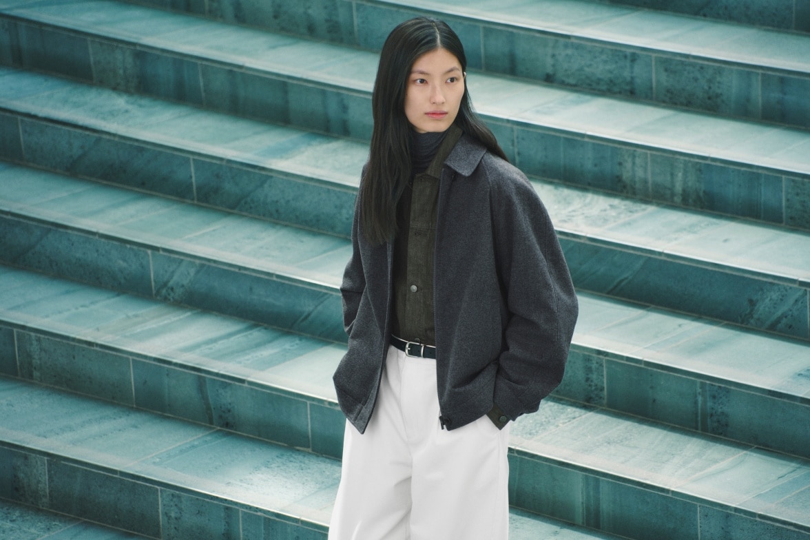 model wearing dark grey jacket over a green shirt and white trousers with a black belt while walking down some green tiled steps