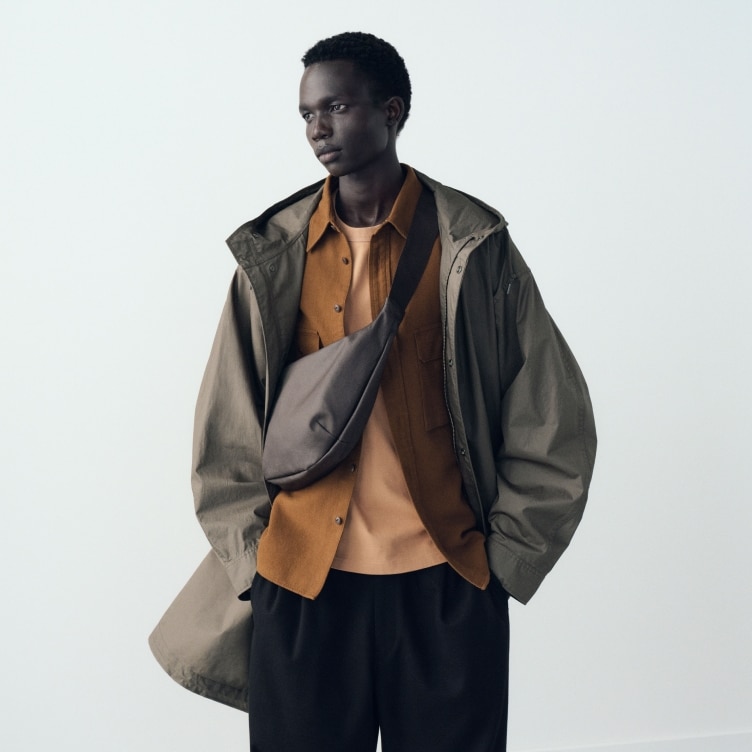 model wearing a dark beige coat over a rust coloured overshirt and pale orange T-shirt  and black trousers with a dark brown cross body bag