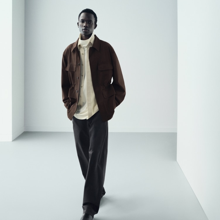 model wearing a brown overshirt over an off white open shirt and white turtleneck with dark grey trousers and black shoes