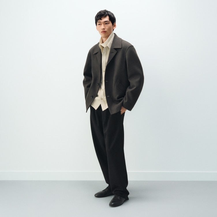 model wearing dark grey pea coat over an off white shirt and white turtleneck with black trousers and shoes
