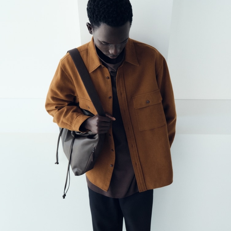 model wearing rust coloured over shirt with a dark brown top, black trousers and a dark brown shoulder bag