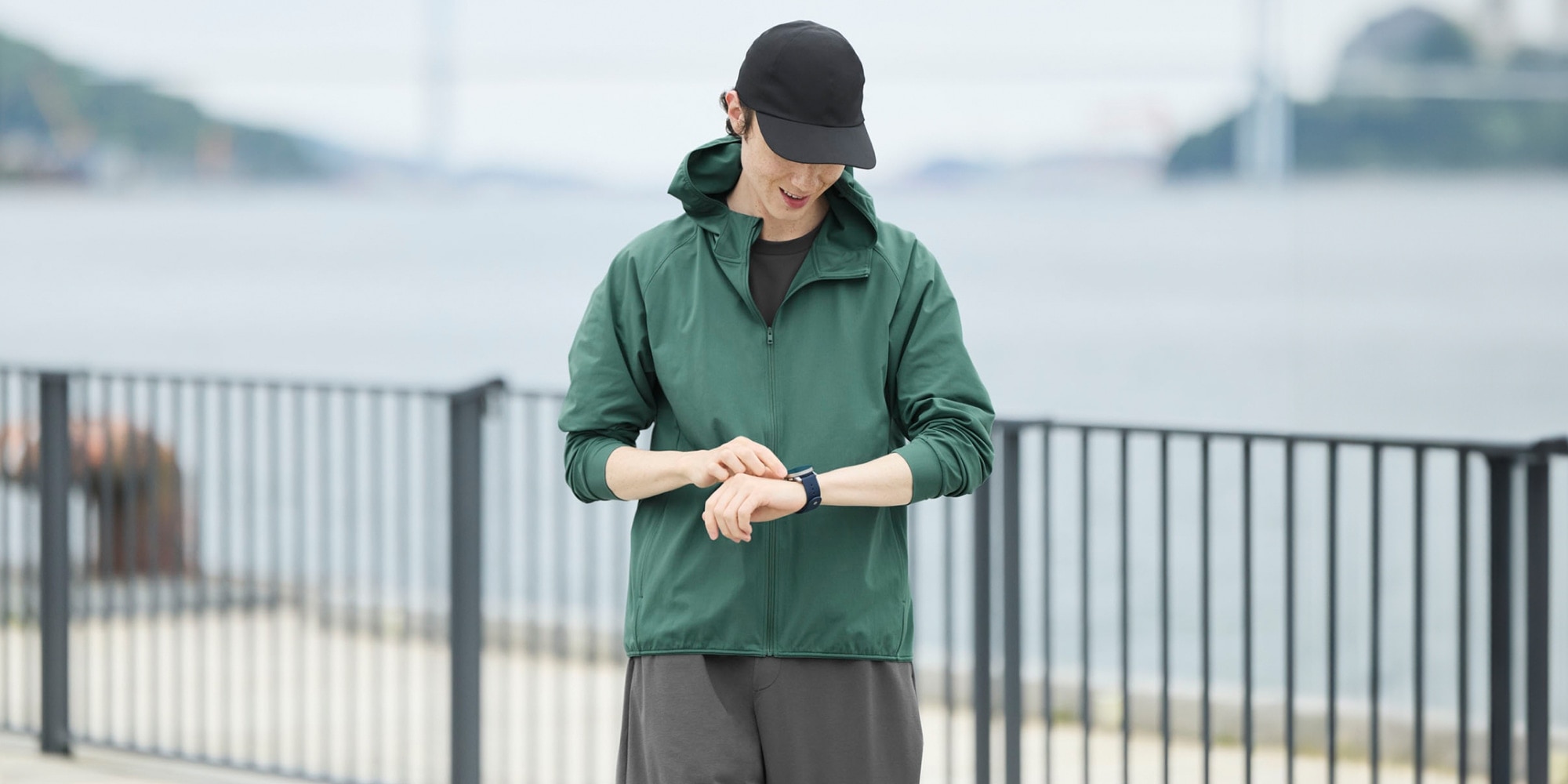 model wearing 2-Way Stretch Cap in black, green jacket and grey joggers while outside by the water