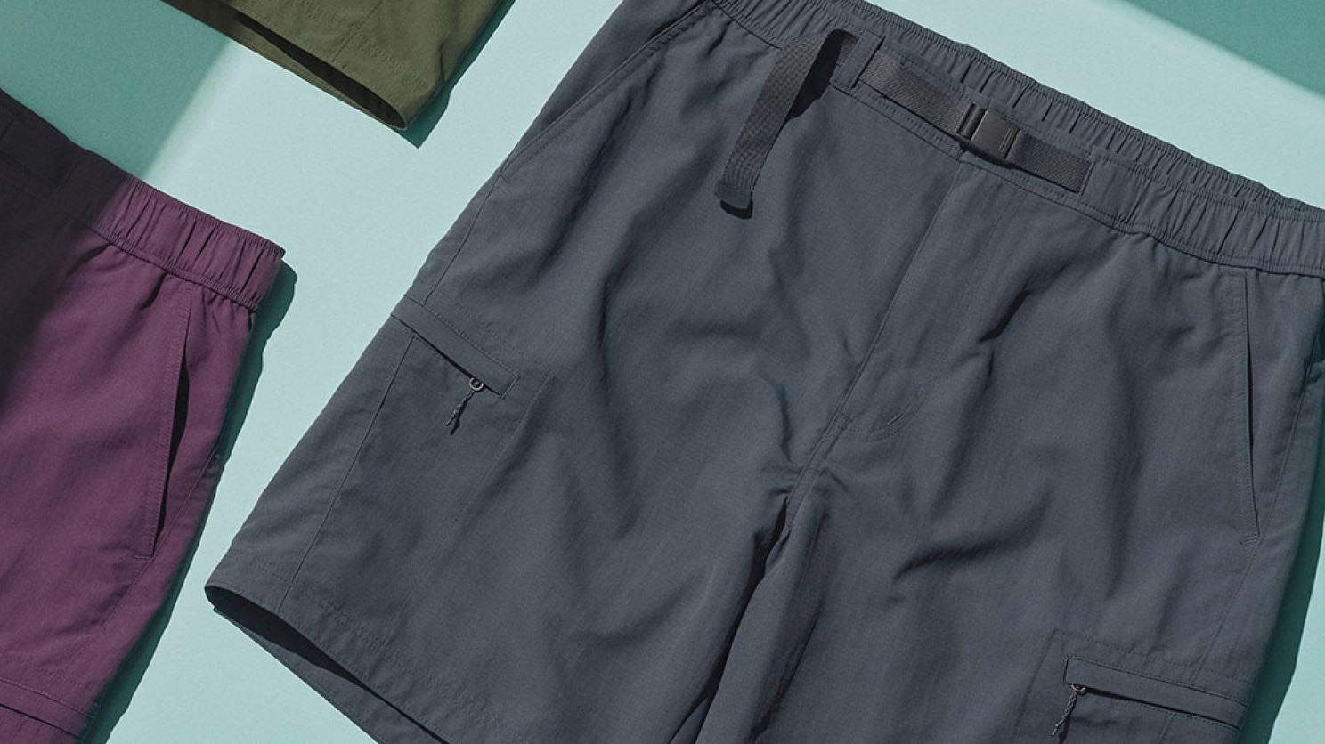 trois paires de short en vert olive, marron et gris foncé, mis à plat sur une surface bleu clair