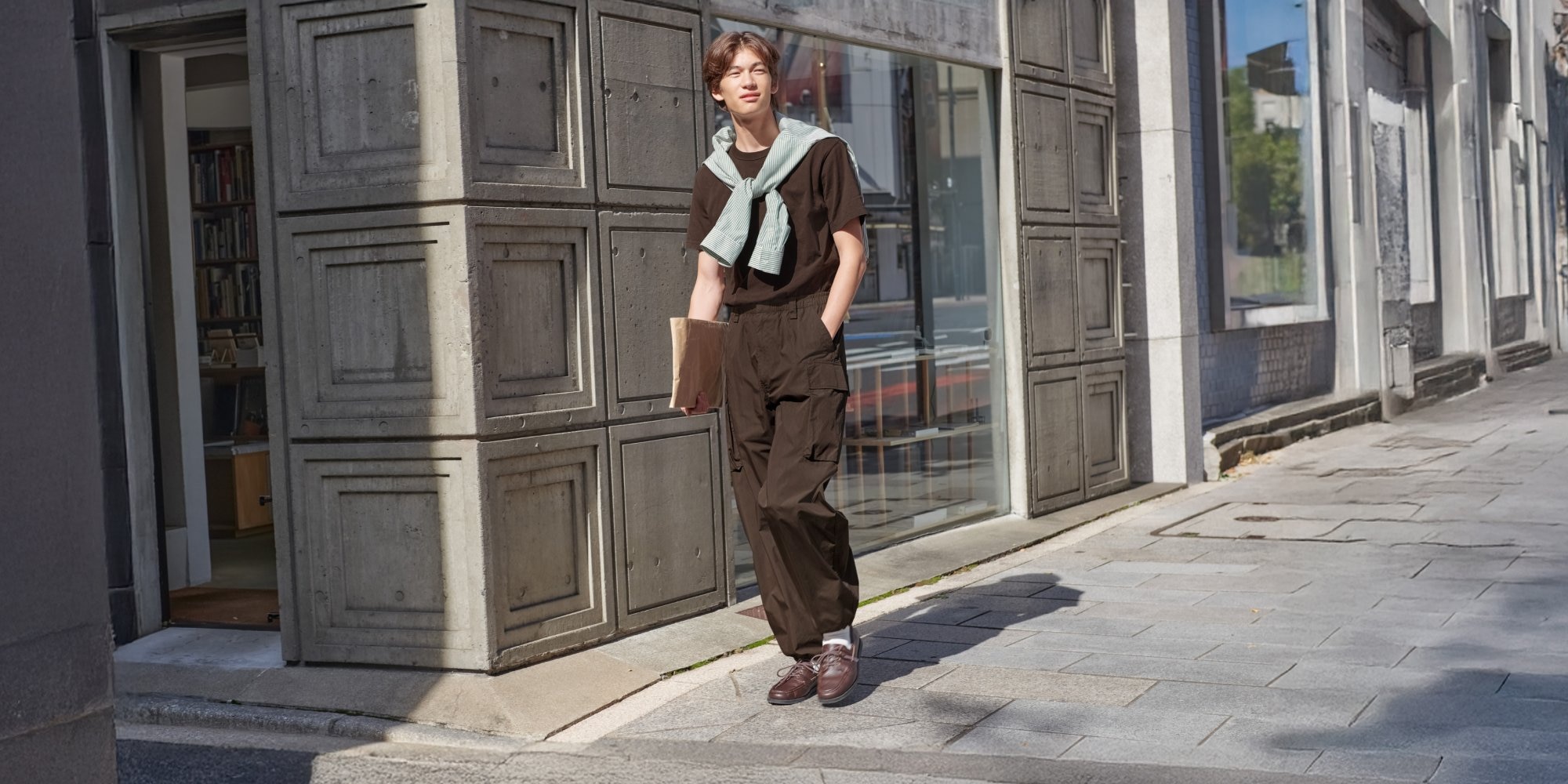 model wearing brown T-shirt, cargo trousers and trainers with a light green striped shirt tied around the shoulders