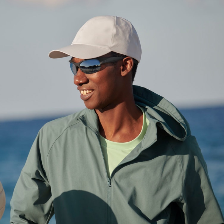 model wearing a white cap, sunglasses, green jacket and light green T-shirt, holding a ball with the sea in the background