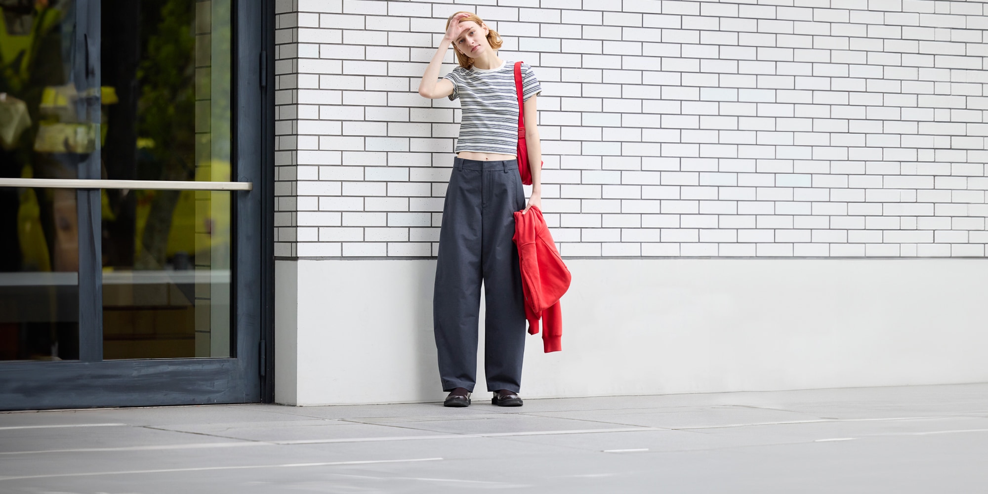 model wearing dark grey barrel leg trousers with a grey and white striped T-shirt and red bag, holding a red hoodie