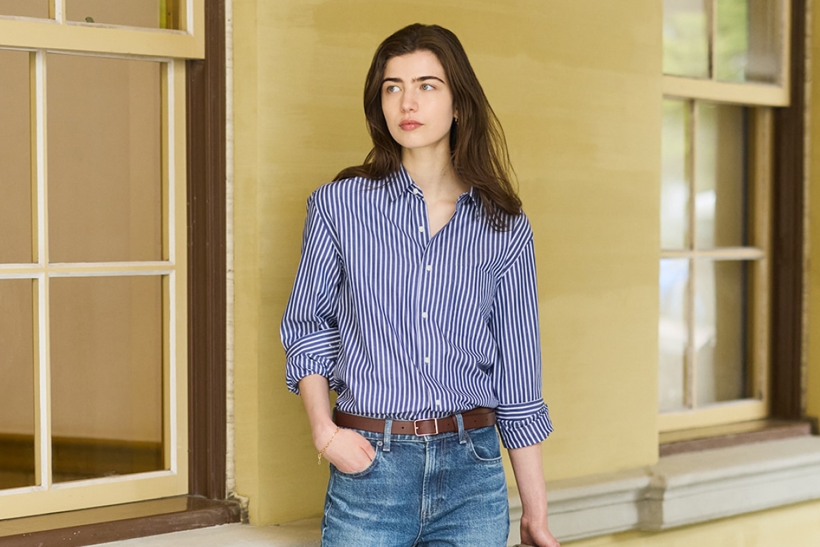 model wearing blue and white striped shirt with mid-wash faded denim jeans and a brown belt while standing in front of a building