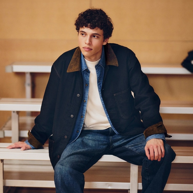 model wearing Coverall over a light wash denim shirt, white T-shirt and mid wash blue denim jeans while sat in a sports hall