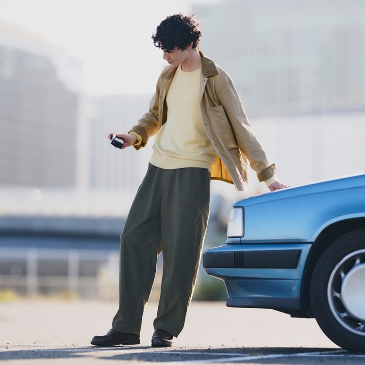 model wearing beige jacket over a yellow crew neck with olive trousers and black shoes while leaning against a blue car