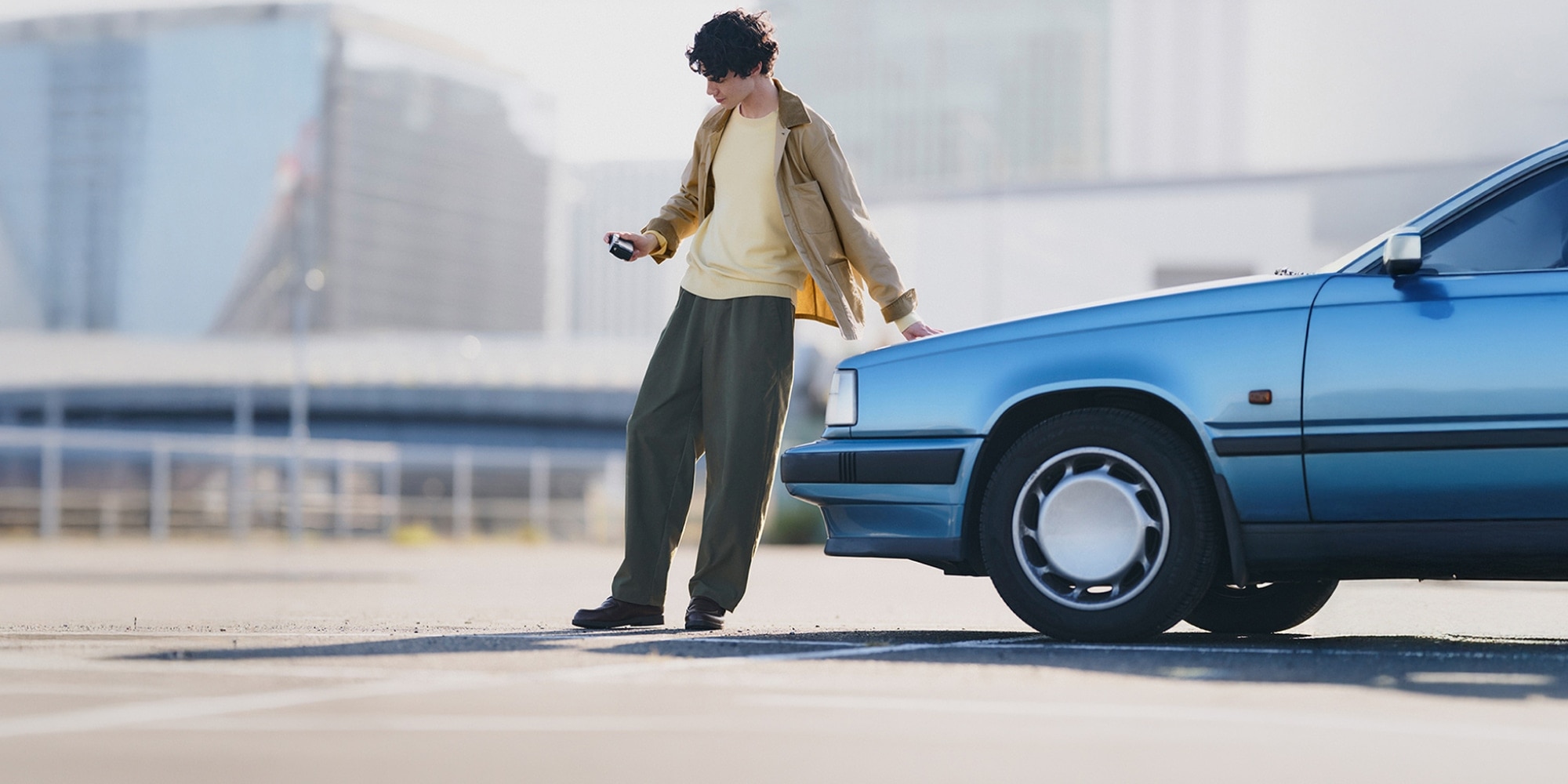 model wearing beige jacket over a yellow crew neck with olive trousers and black shoes while leaning against a blue car