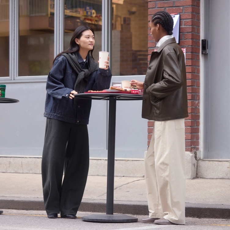 two models wearing jackets and barrel leg trousers while leaning on an outside table with pizza and drinks in takeaway cups