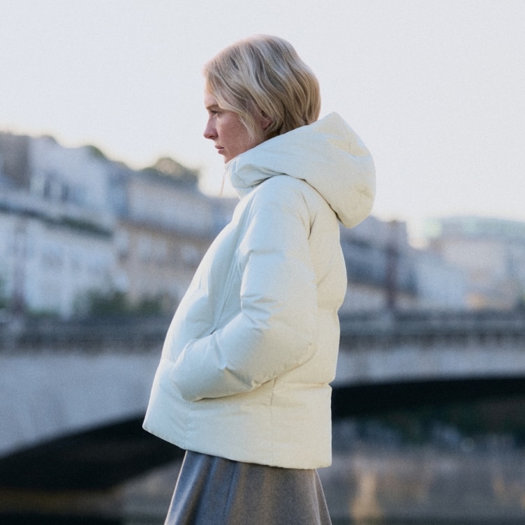 model wearing Seamless Down Parka in off white with a grey skirt with a bridge in the background
