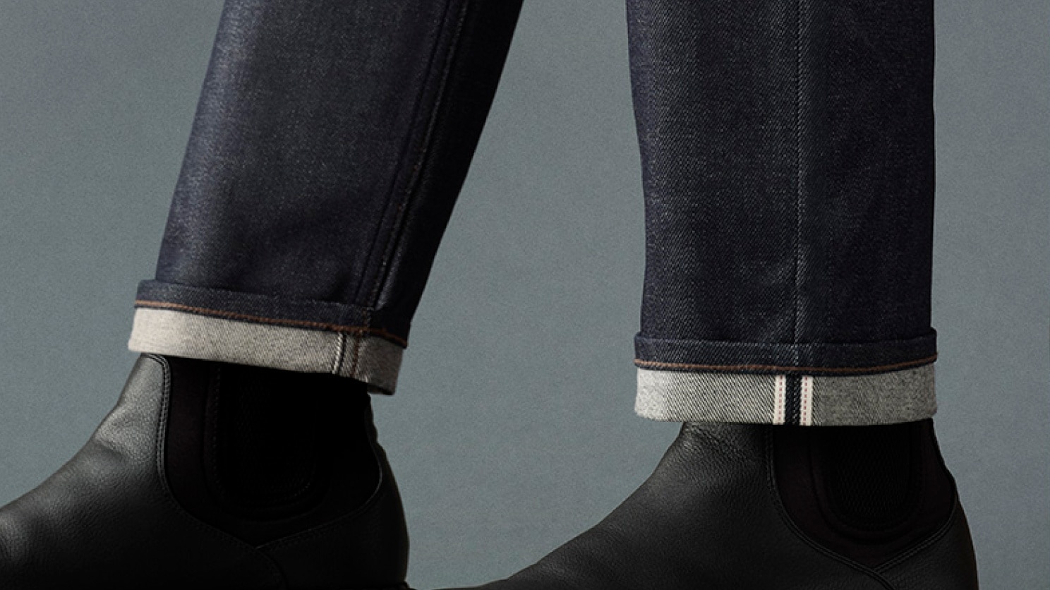 model wearing Selvedge Slim Straight Jeans in dark wash denim with black chelsea boots and a light grey shirt while sitting on a brown wooden chair