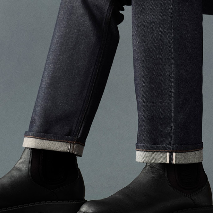 Model draagt ​​een Selvedge Slim Straight jeans in een donkere denimwassing, zwarte Chelsea boots en een lichtgrijs shirt, terwijl hij op een bruine houten stoel zit.