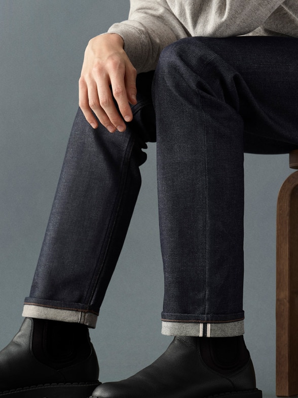 model wearing Selvedge Slim Straight Jeans in dark wash denim with black chelsea boots and a light grey shirt while sitting on a brown wooden chair