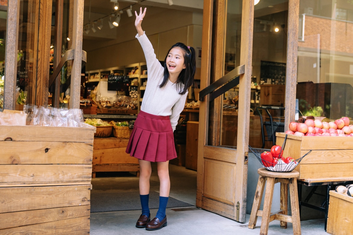 model wearing Ribbed Crew Neck T-Shirt in light grey with a red pleated skort, blue socks and brown loafers while waving outside a grocery store