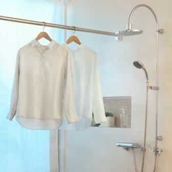 two linen shirts in beige and white hanging on a shower curtain pole in a bathroom