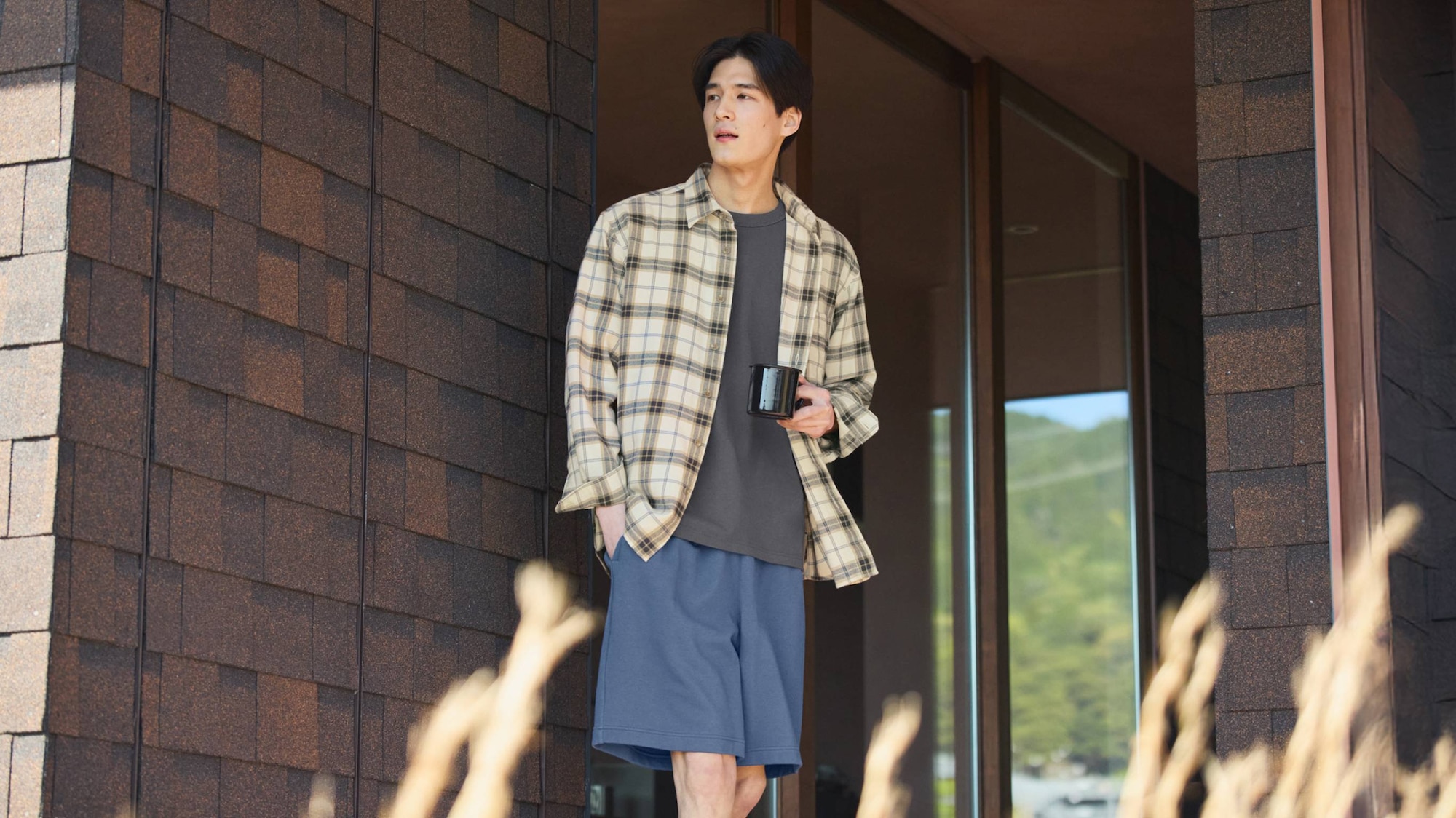 model wearing an off white checked flannel shirt over a dark grey T-shirt and blue trousers holding a mug