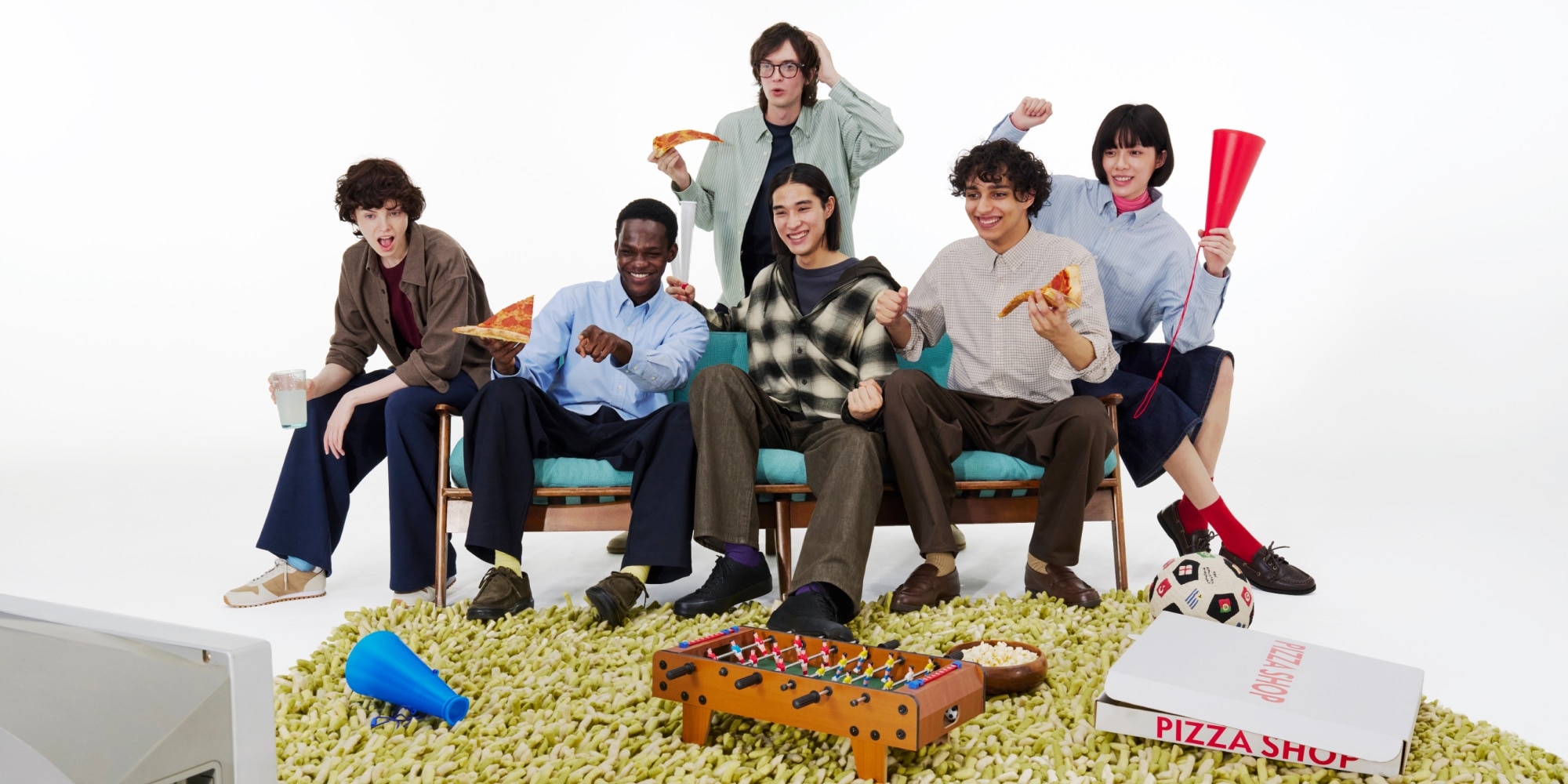 Group wearing casual layered outfits, including lightweight shirts, relaxed-fit pants, and everyday essentials, gathered indoors with pizza and games.