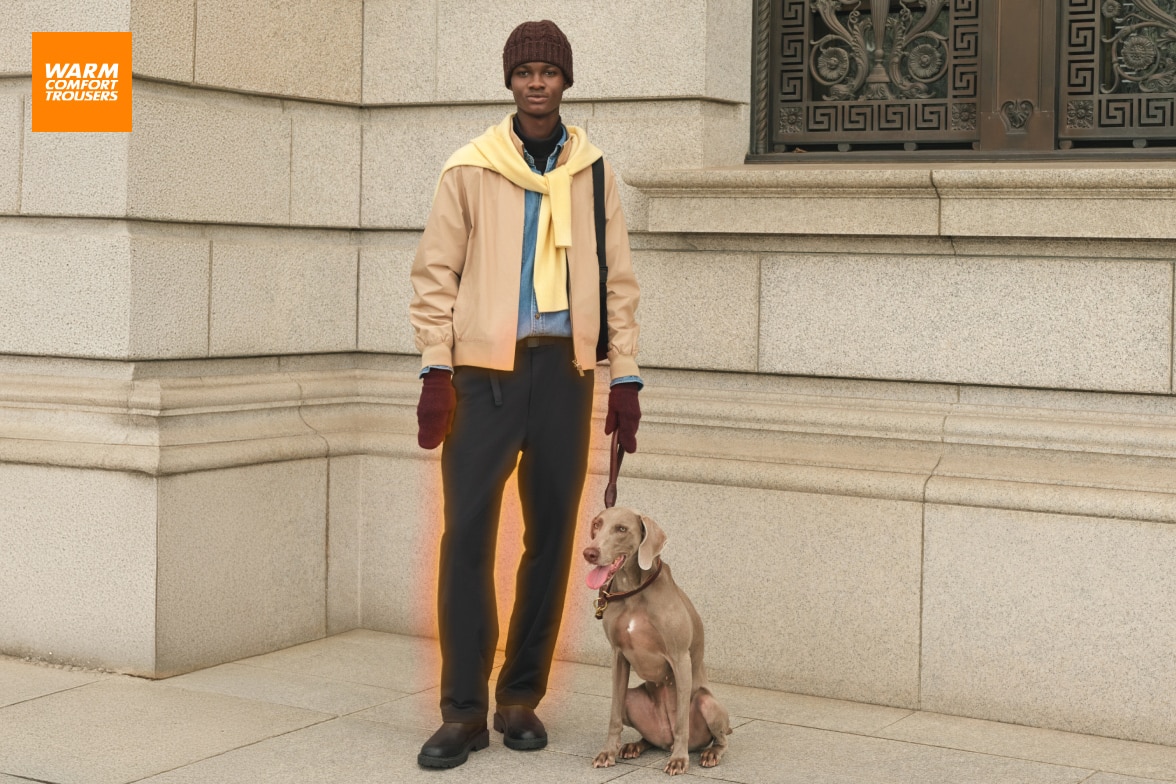 model wearing warm lined trousers in dark grey with a denim shirt, beige jacket and red hat and mittens, with a pale yellow jumper draped over the shoulders while standing with a Weimaraner dog on a lead