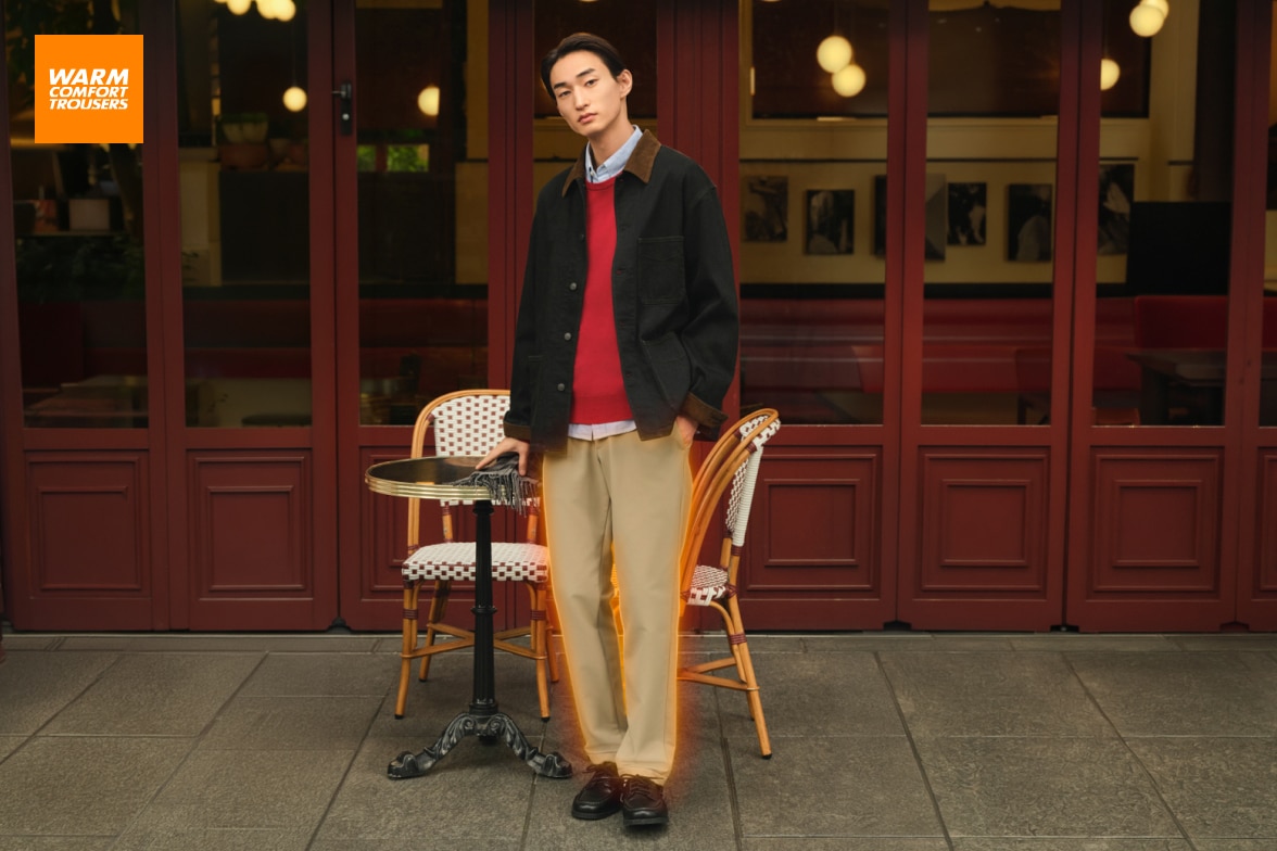 model wearing warm stretch trousers in beige with a light blue shirt, red jumper and denim jacket while stood outside a café