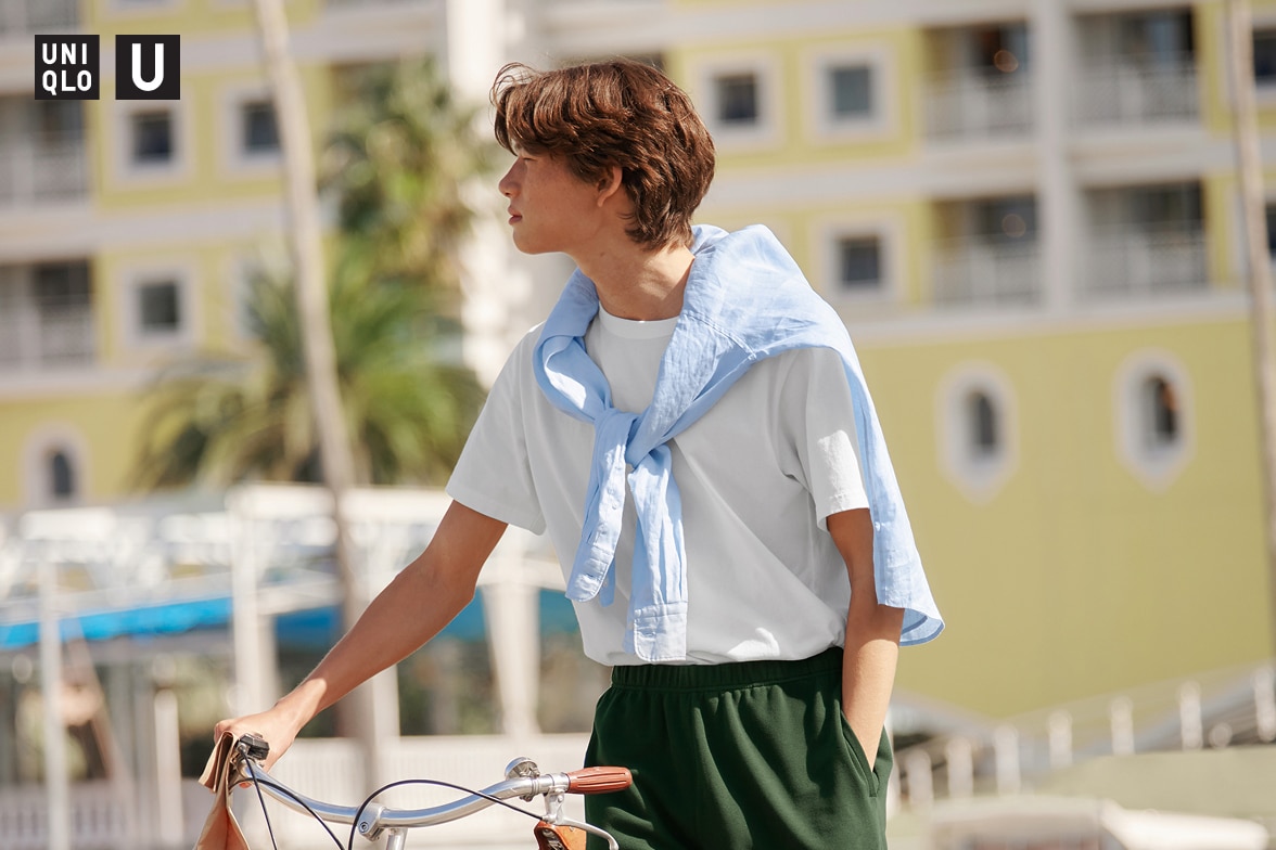 model wearing Crew Neck T-Shirt in white with dark green shorts and a light blue shirt tied around the shoulders while pushing a bike
