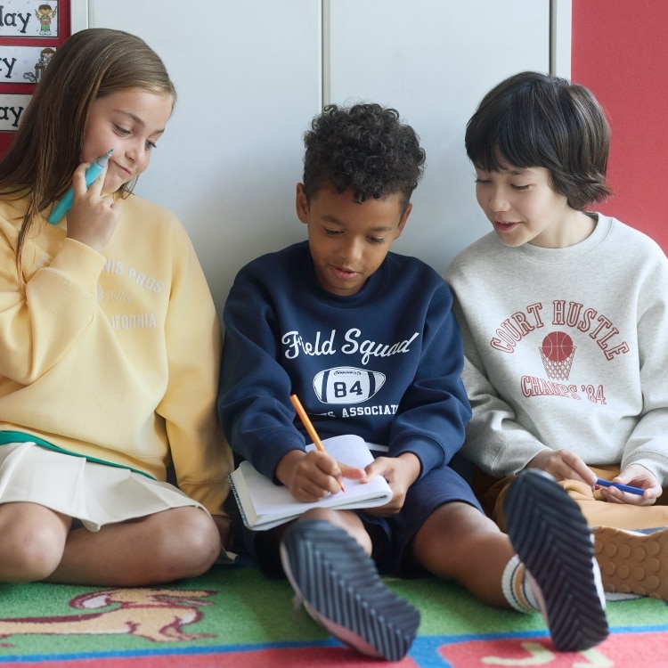 Three kids sitting together writing and drawing, wearing sweatshirts and casual clothes.