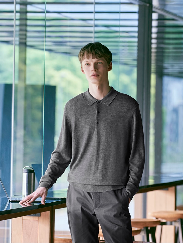 model wearing grey knitted polo with grey trousers, next to a window seat with a laptop