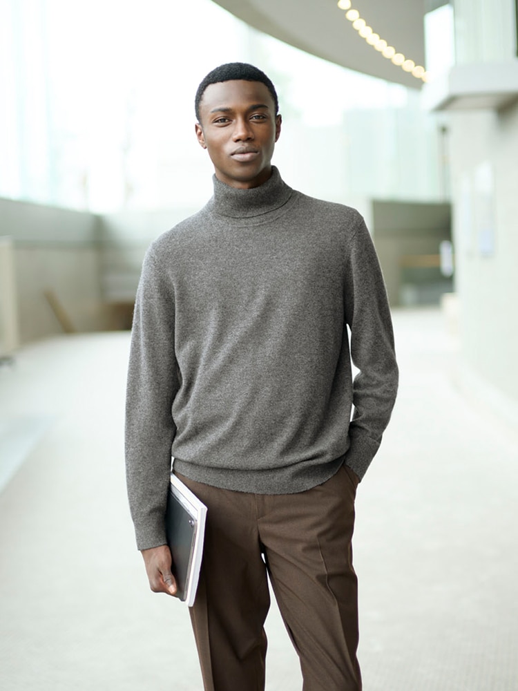 model wearing a grey turtleneck jumper with brown trousers, while holding a tablet and some paper