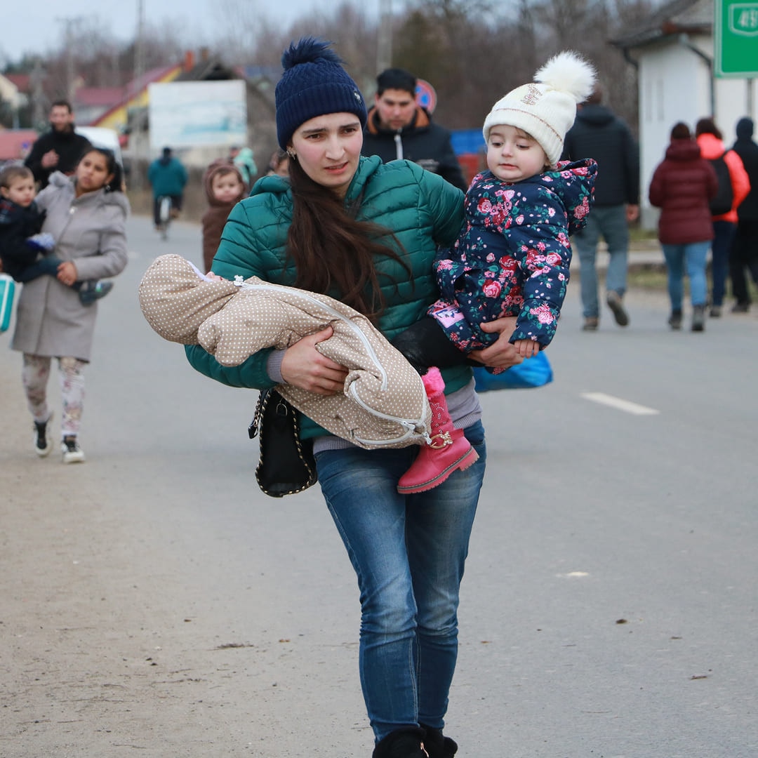 Woman carrying two young children along a road with other families on a cold day
