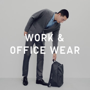 model wearing a gray blazer with matching gray dress pants and a blue stand collar shirt. Leaning towards a backpack.