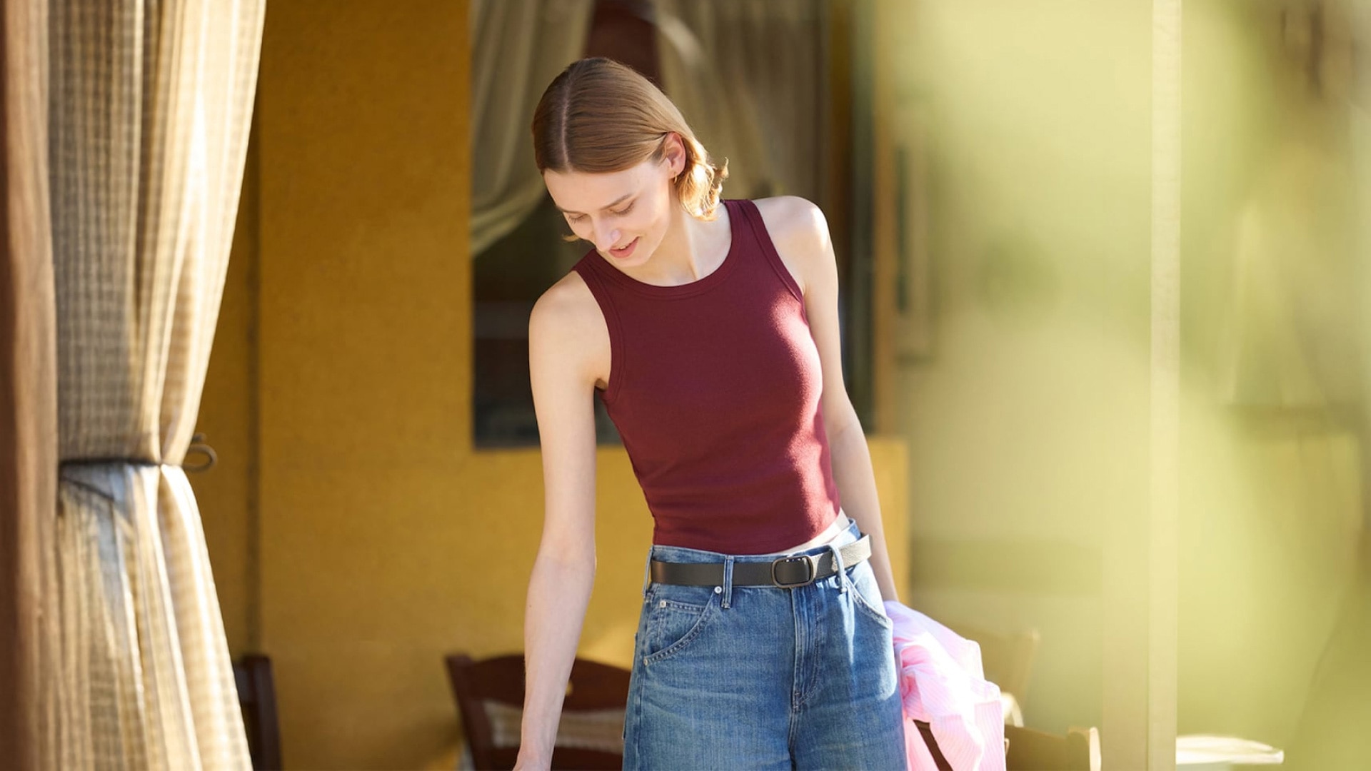 Model wearing maroon bra top, black belt, and denim pants, while holding a pink linen shirt.