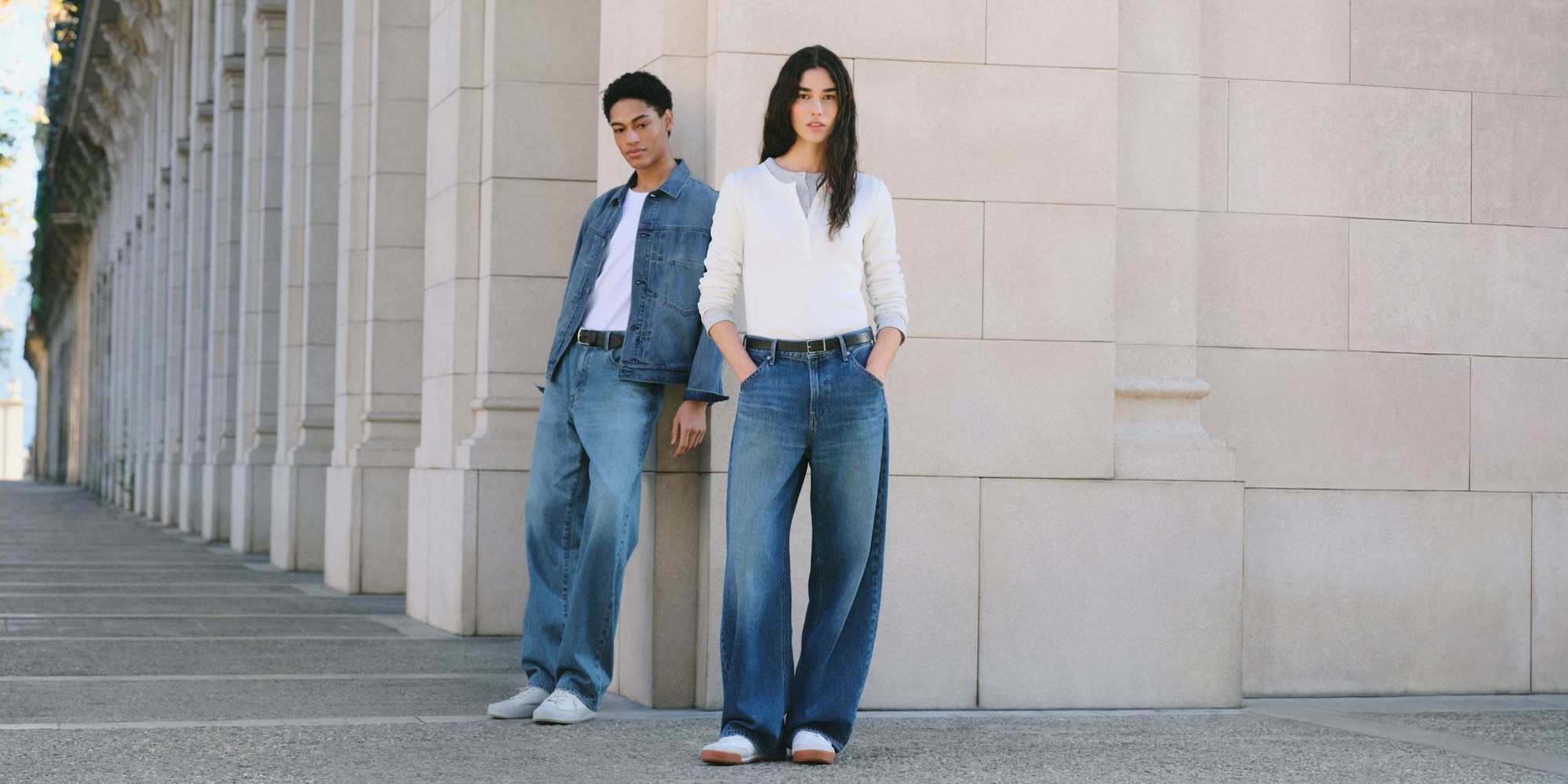 Two models wearing straight and wide denim jeans. the man on the left is wearing a denim jacket and white tee while the woman at the front is wearing a white long sleeve henley over a gray cardigan. Both models are wearing black classic belts.