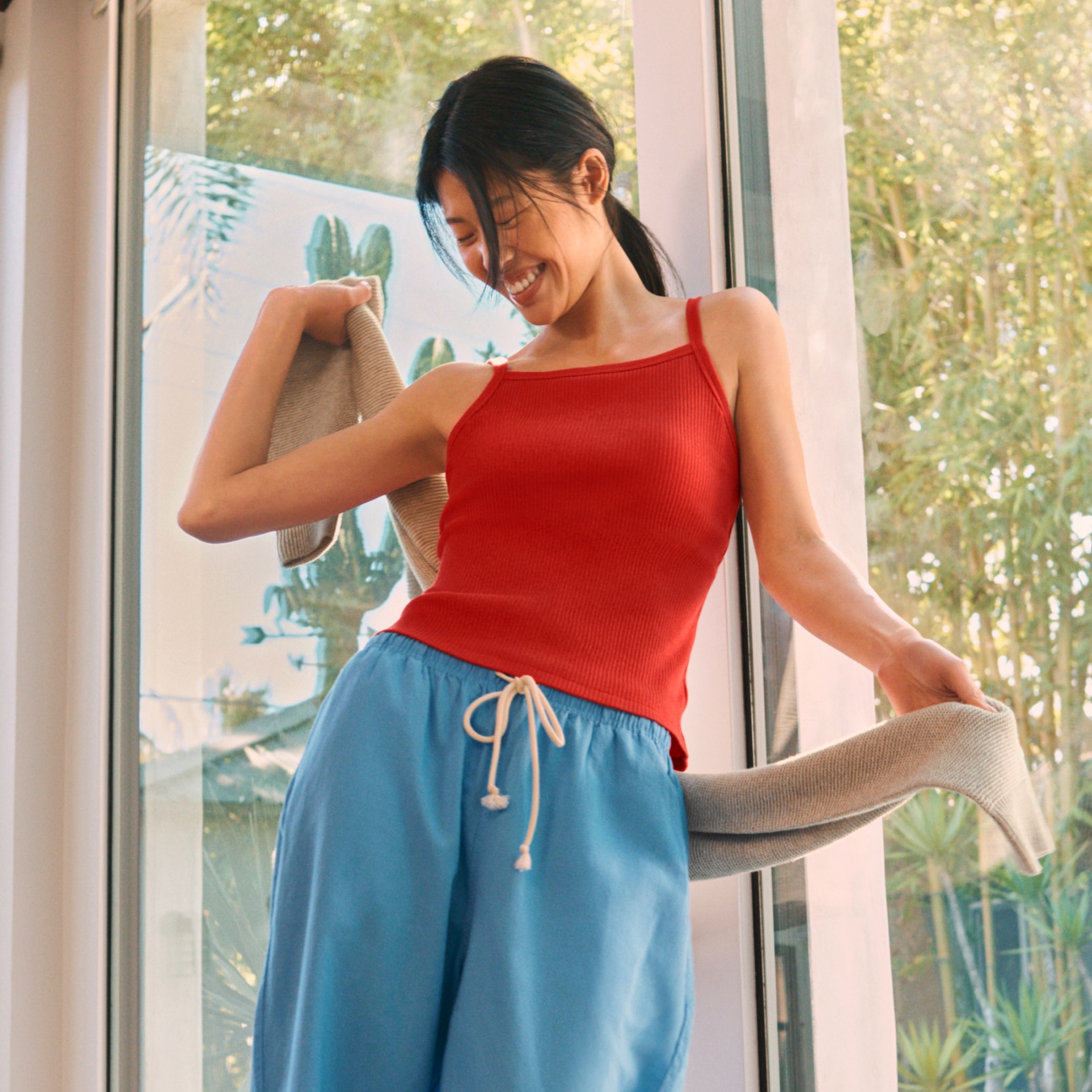 Woman wearing red square neck bra camisole, blue pull on pants, and holding a knit beige sweater