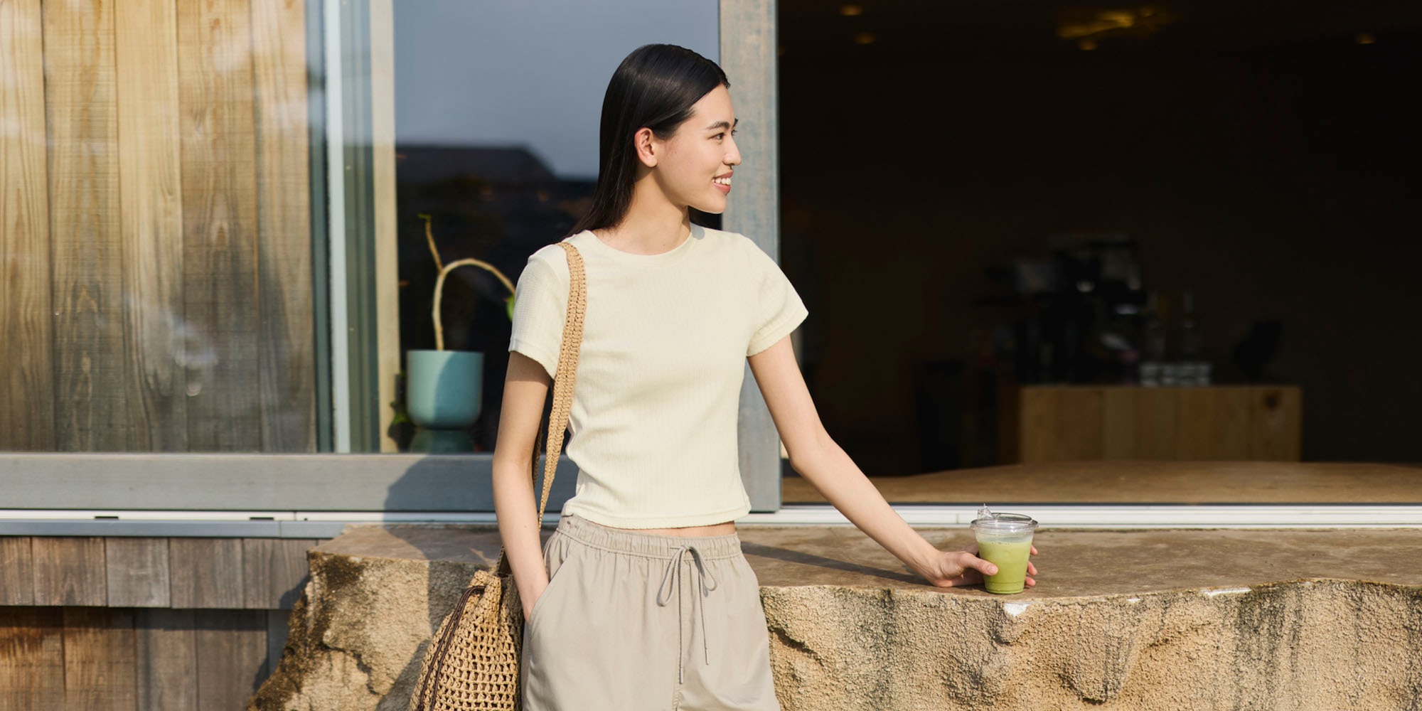 Woman outside wearing a beige short sleeve shirt, pull on brown pants, and a crochet shoulder bag with matcha in hand